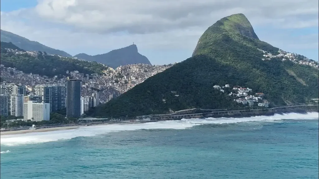 Vista aérea da Praia de São Conrado com ondas fortes, o Morro Dois Irmãos ao fundo e o Cristo Redentor visível no horizonte.