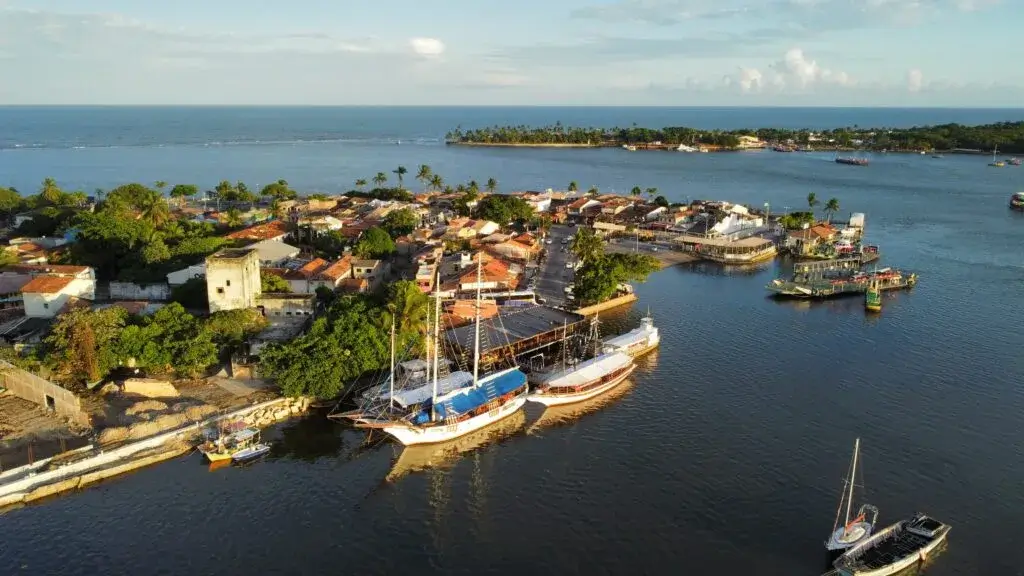 Vista aérea do centro histórico e do litoral de Porto Seguro com barcos ancorados no mar tranquilo, casinhas coloridas e vegetação tropical ao redor, destacando a região próxima à Passarela do Descobrimento e o encontro entre cidade e oceano.