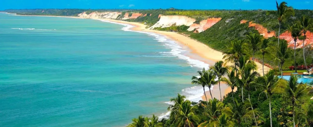 Vista panorâmica da Praia de Pitinga com falésias coloridas em tons de vermelho e branco, mar azul-turquesa calmo e faixa de areia dourada cercada por coqueiros tropicais, um dos cenários mais paradisíacos próximos a Porto Seguro.