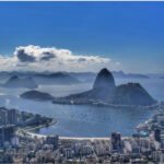 Vista aérea do Pão de Açúcar e da Baía de Guanabara com mar azul, barcos ancorados e a orla urbana do Rio de Janeiro ao fundo em dia ensolarado.